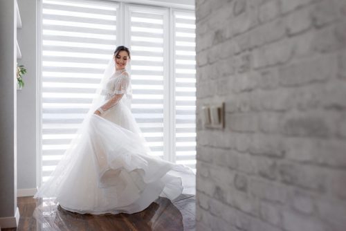smiled pretty bride is turning around room near white brick wall dressed fashionable dress wedding fashion scaled e1628567673966